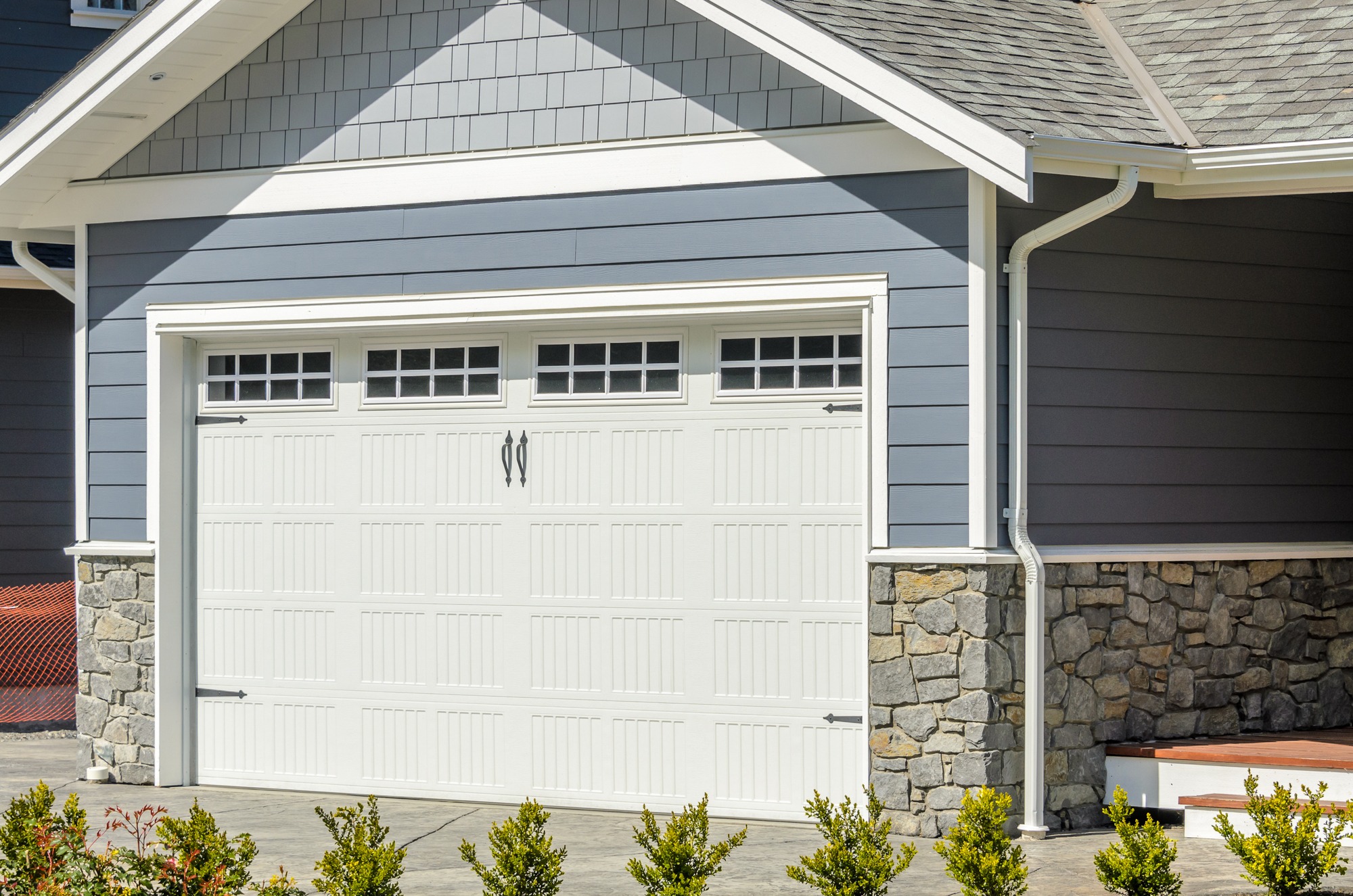 Stylish gray and white house exterior featuring a single garage door with windows, complemented by stone accents and leafy shrubs.