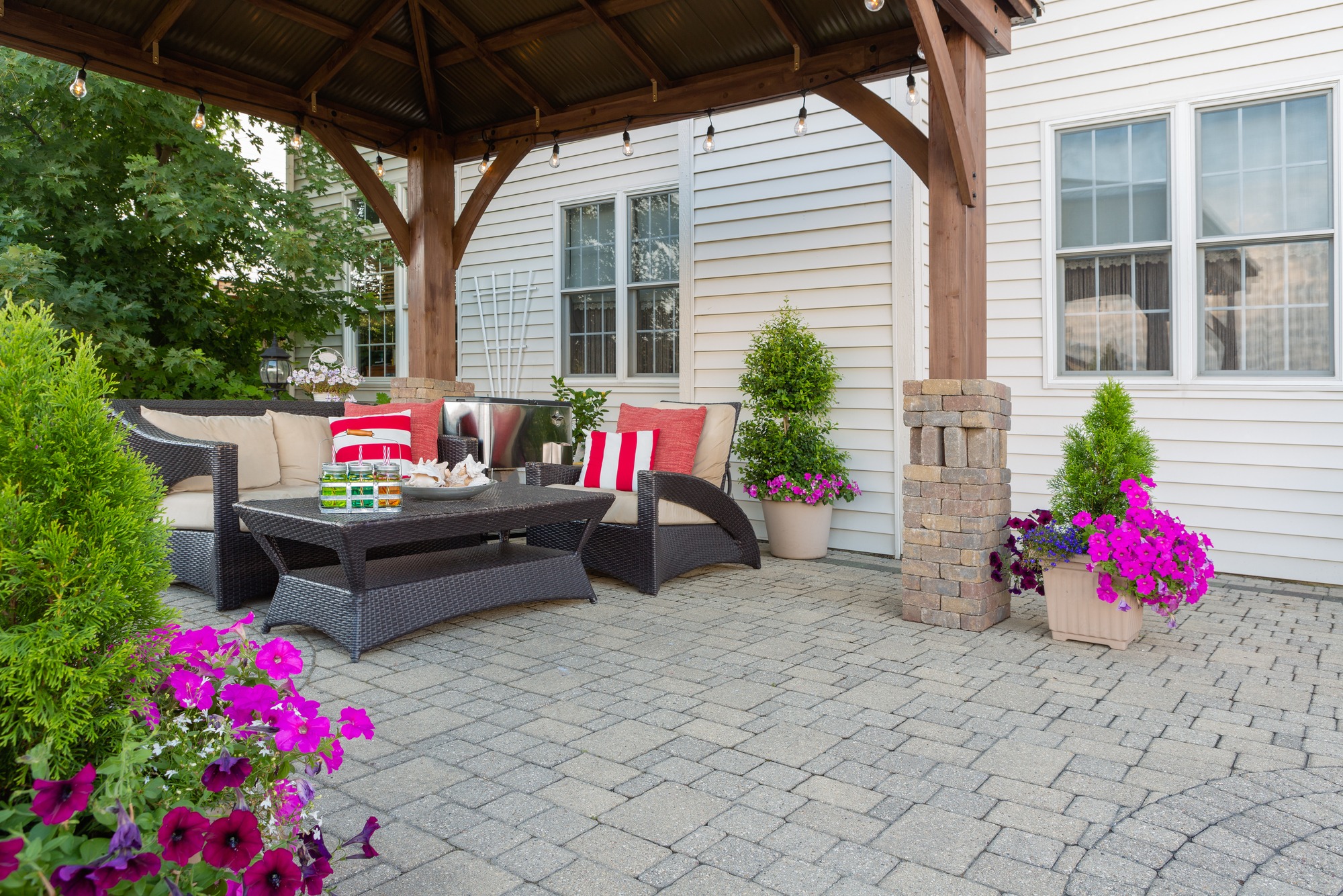 A cozy outdoor seating area under a wooden gazebo, featuring plush sofas with red and white cushions, a coffee table with snacks, and potted flowers.